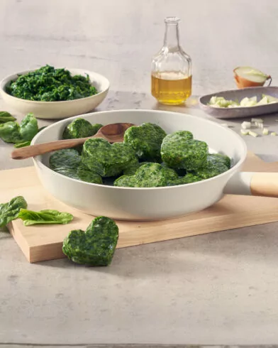 Na desce patelnia z pierogami ze szpinakiem FRoSTA; obok świeży szpinak, cebula, mrożony szpinak FRoSTA, pieprz i oliwa.