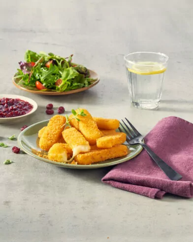 Talerz panierowanych paluszków serowych FRoSTA z sosem żurawinowym, sałatą, wodą z cytryną i fioletową serwetką na szarym tle.