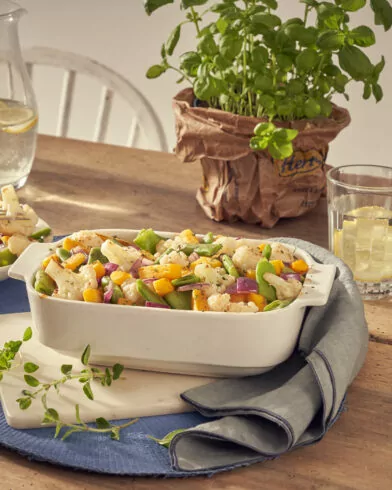 Na stole stoi ceramiczne naczynie z sałatką z warzyw FRoSTA, obok woda z cytryną, dzbanek, zioła i dodatkowa miska sałatki.