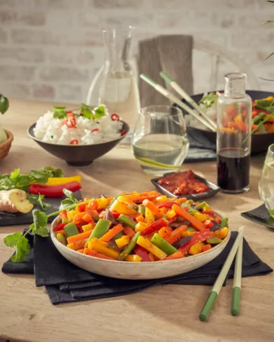 Miska kolorowych warzyw na patelnię FRoSTA na stole z ryżem, sosem sojowym i świeżymi dodatkami, idealny pomysł na podanie.