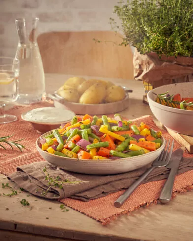 Drewniany stół z warzywami na patelnię FRoSTA, ziemniakami, sosem, wodą i ziołami w tle; idealne podanie.