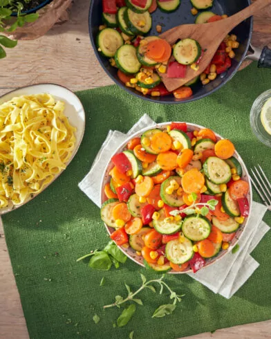 Nakrycie stołu: talerz żółtego makaronu, warzywa na patelnię FRoSTA, sałatka, woda z cytryną i świeże zioła.