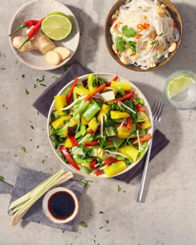 Miska kolorowych warzyw azjatyckich FRoSTA z papryką i groszkiem, obok makaron, imbir, chili, limonka oraz sos sojowy.