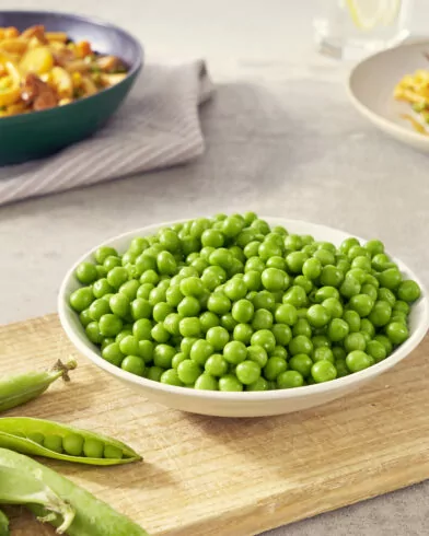 Miska groszku zielonego FRoSTA na desce, obok strąki; w tle dwa talerze makaronu, jeden z sosem, drugi z groszkiem FRoSTA.