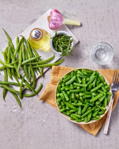 Miska pokrojonej fasolki szparagowej FRoSTA na serwetce z widelcem, obok całej fasolki FRoSTA, czosnek, oliwa i zioła.