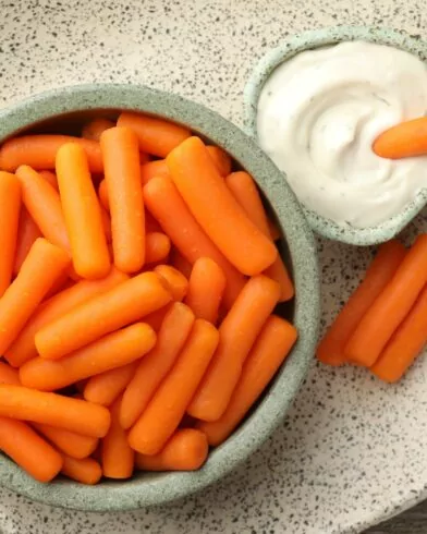 Miska mini marchewki FRoSTA obok miseczki białego dipu, kilka marchewek FRoSTA między nimi, wszystko na nakrapianym talerzu.