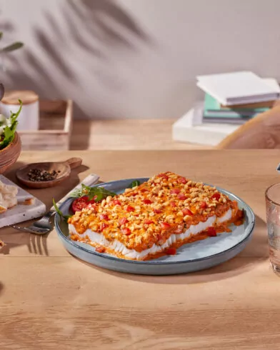 Drewniany stół z sałatką, bagietką i pieczoną fetą z pomidorami FRoSTA, szklanką wody i karafką z cytryną w słońcu.