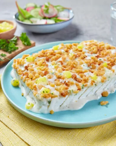 Warstwowa sałatka z ziemniakami FRoSTA, posypana krakersami i szczypiorkiem, na niebieskim talerzu z żółtą serwetką.