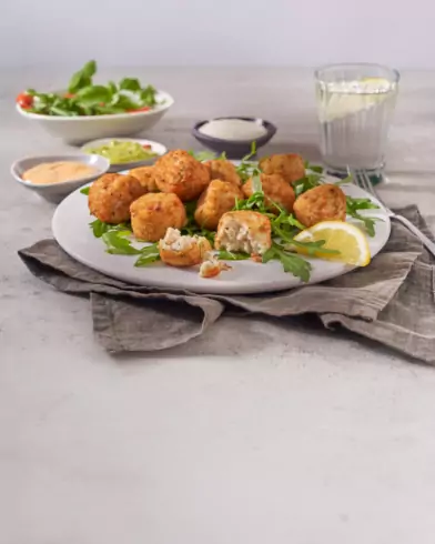 Talerz złotych klopsików rybnych FRoSTA na rukoli z cytryną. W tle sosy, woda i sałatka, wszystko na jasnoszarym tle.